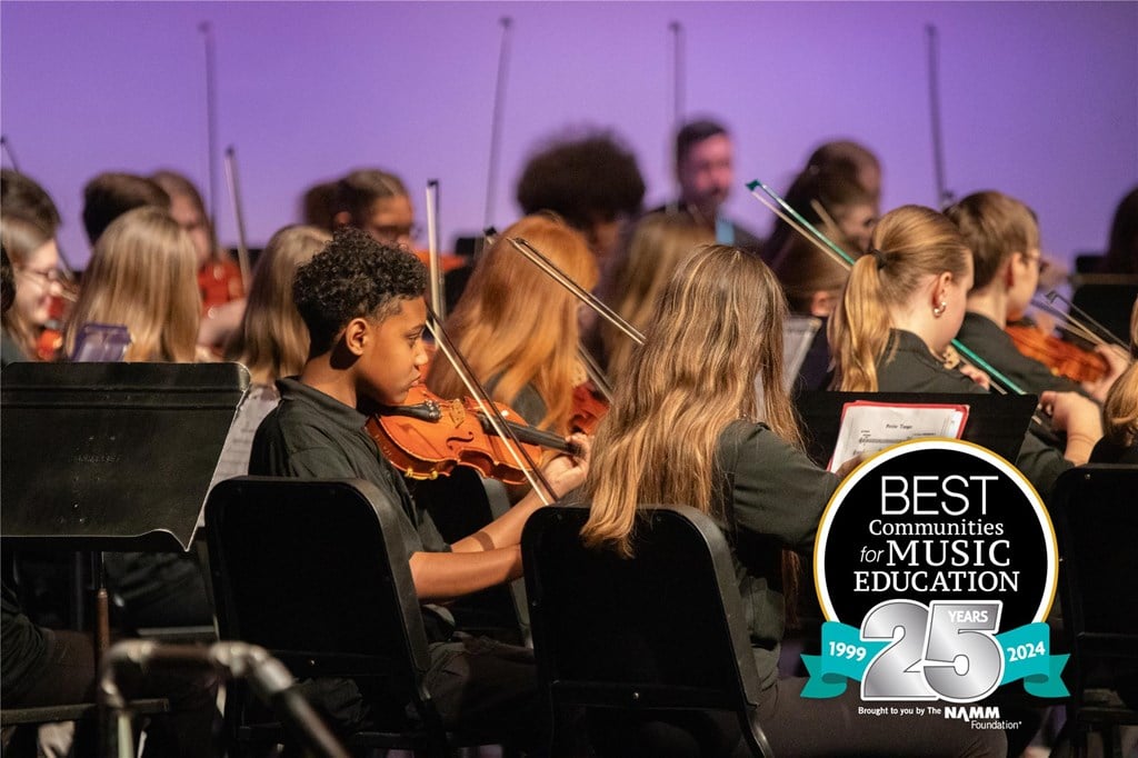 Students playing the violin on stage with the logo for the Best Communities for Music Education's 25th anniversary logo placed in the bottom right-hand corner.